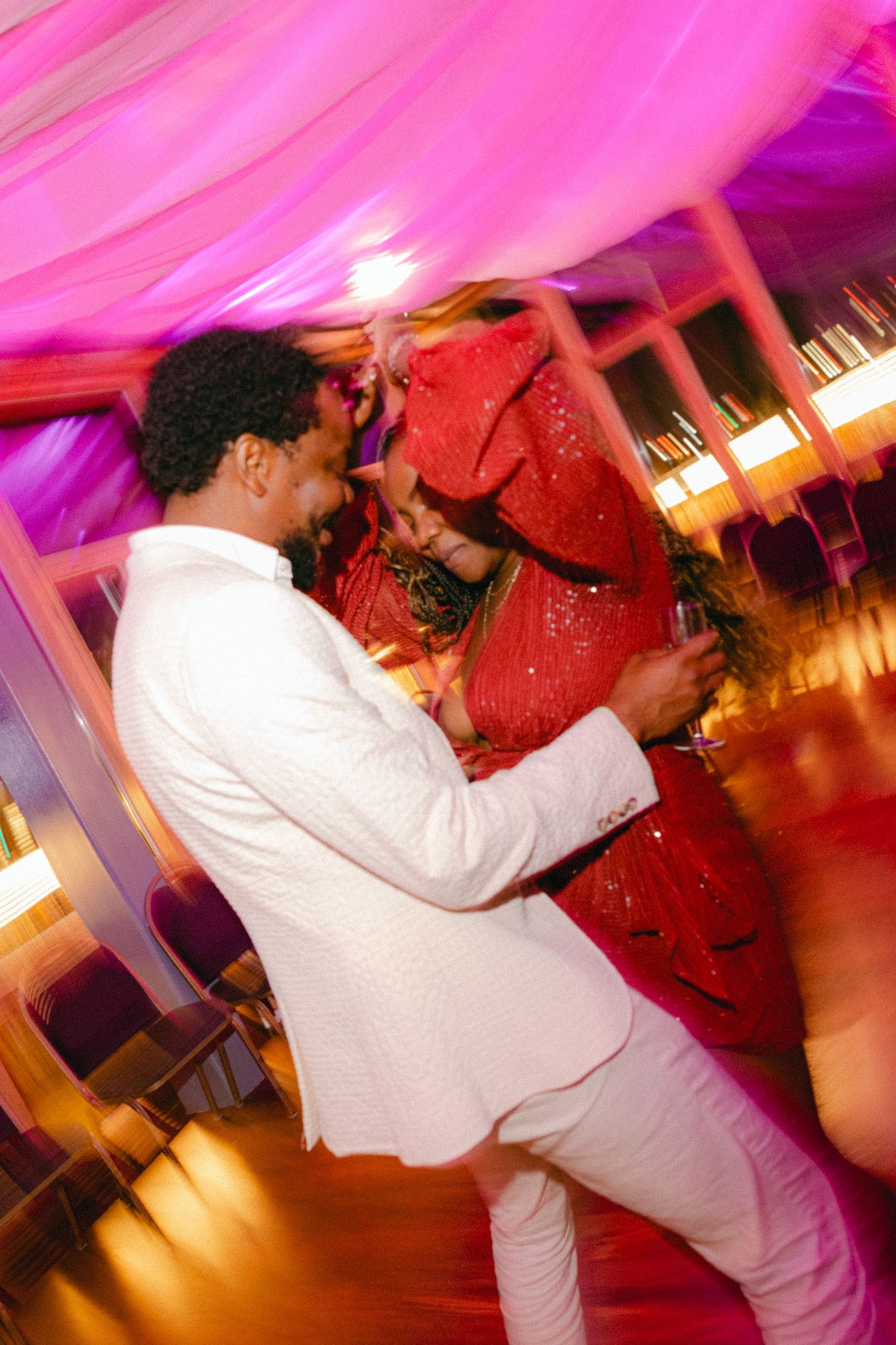 A couple dances intimately under pink and purple lighting at what appears to be a wedding reception or formal event.