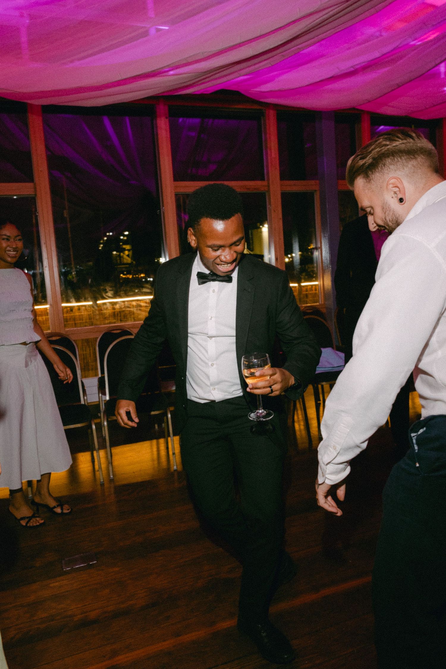 Wedding guests in formal attire dance and celebrate on a wooden dance floor under pink-tinted lighting.