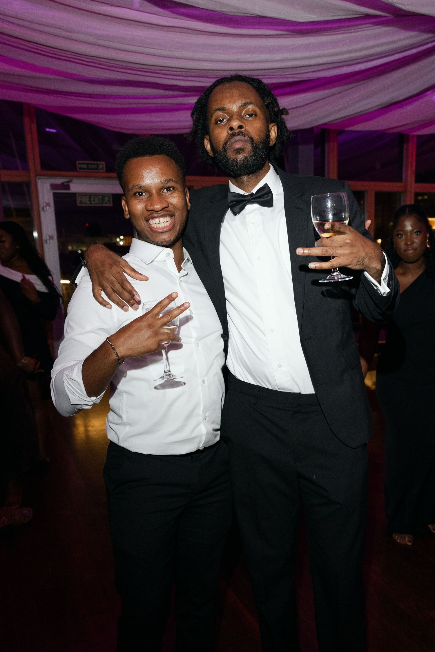 Two people in formal attire share a laugh at a party under purple-lit draped ceiling decor.