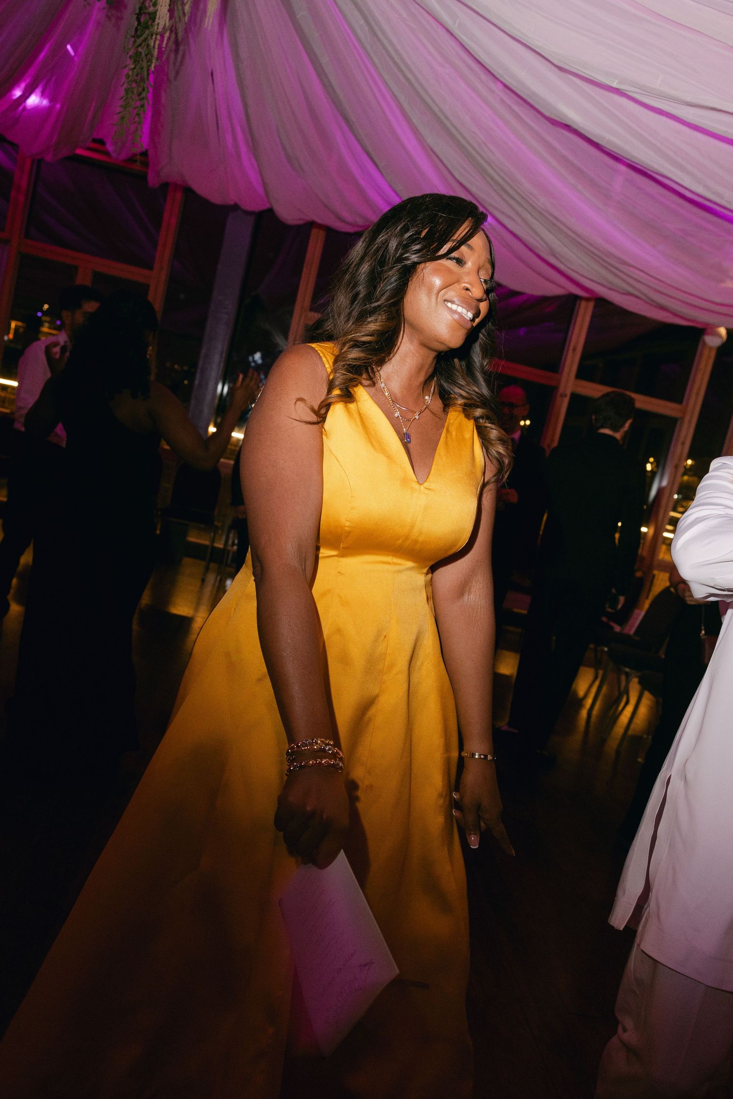 A person in a bright yellow dress dances and smiles at an evening event with purple lighting and draped decor.