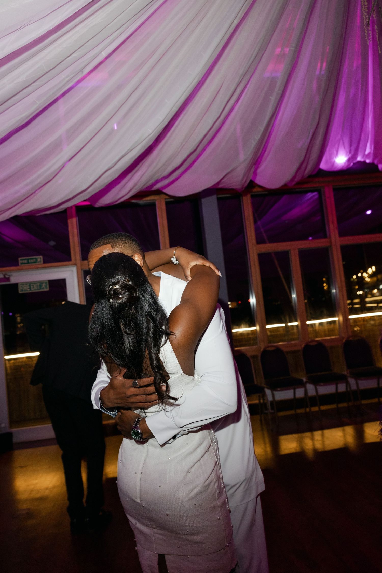 A couple embraces on the dance floor under purple-tinted tent lighting at an evening reception.