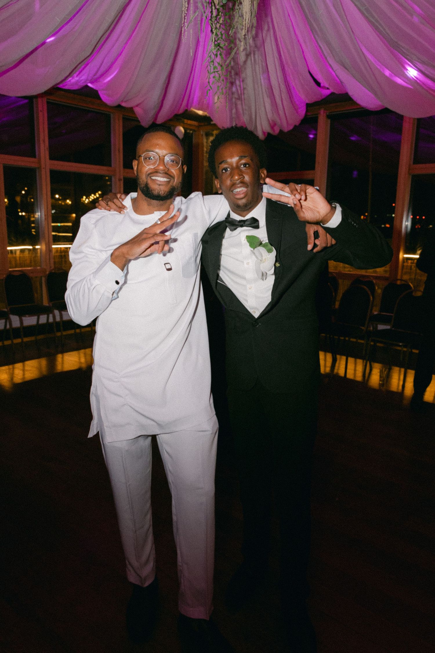 Two people in formal attire, one in white and one in black, posing together at an event with pink lighting.