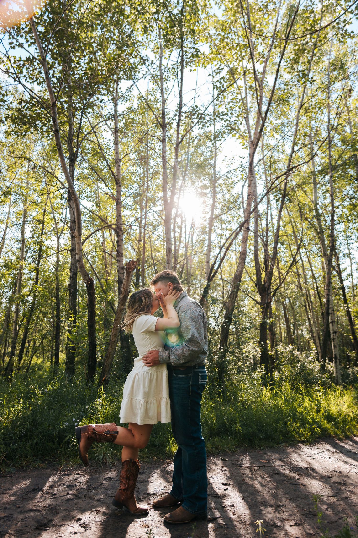 Couple sharing a romantic kiss in a sunlit forest, woman in white dress and brown boots, man in jeans and gray shirt. Parks in Edmonton.
