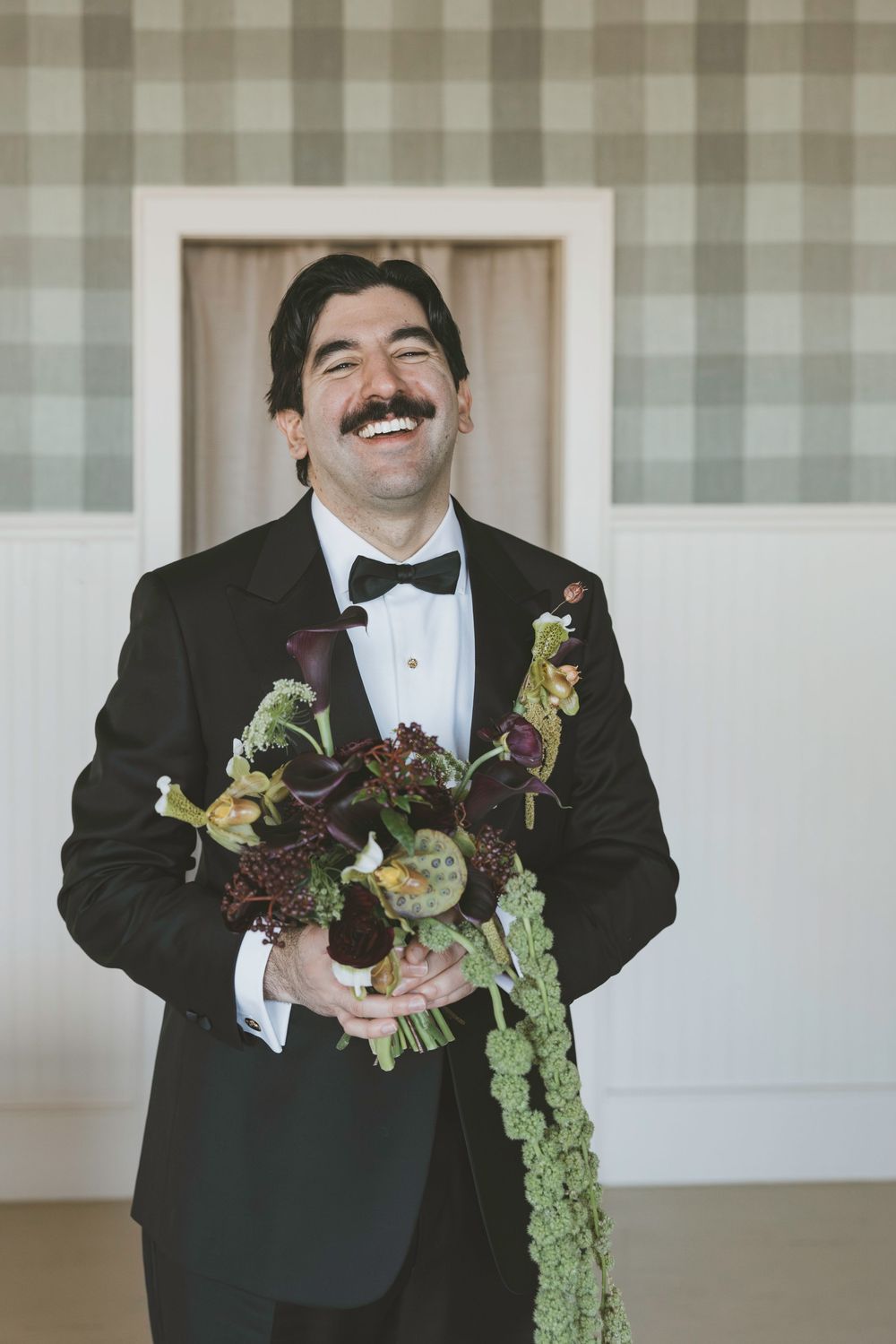 Wedding party members in black tuxedos hold rustic floral bouquets while laughing during a candid moment.