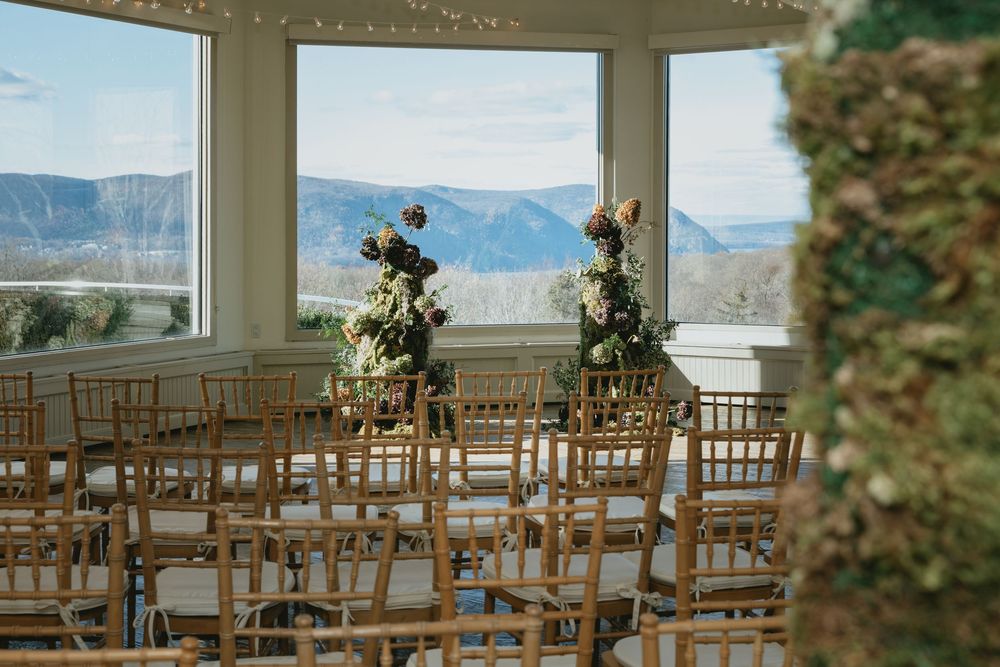 A rustic indoor wedding ceremony setup with wooden chairs facing large windows overlooking mountain views.