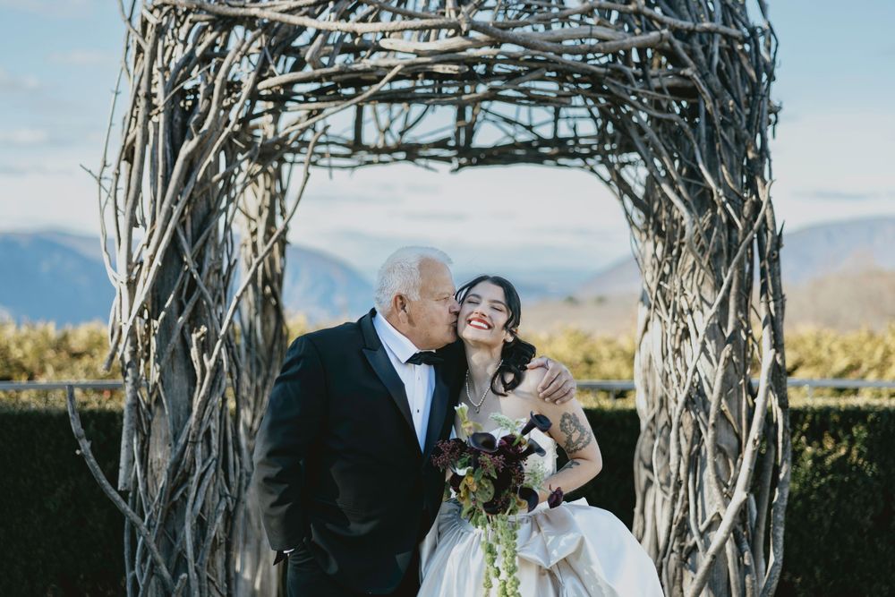 Couple shares romantic moments under rustic wooden arch during outdoor wedding ceremony in scenic mountain setting.