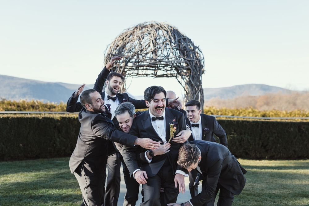 Wedding party laughs together while posing beneath natural woven branch archway at outdoor venue.