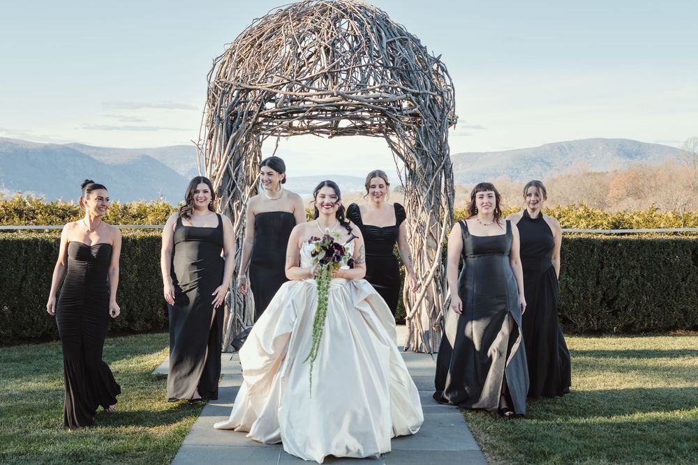 Wedding party in black bridesmaid dresses and white bridal gown pose outdoors against mountain backdrop.