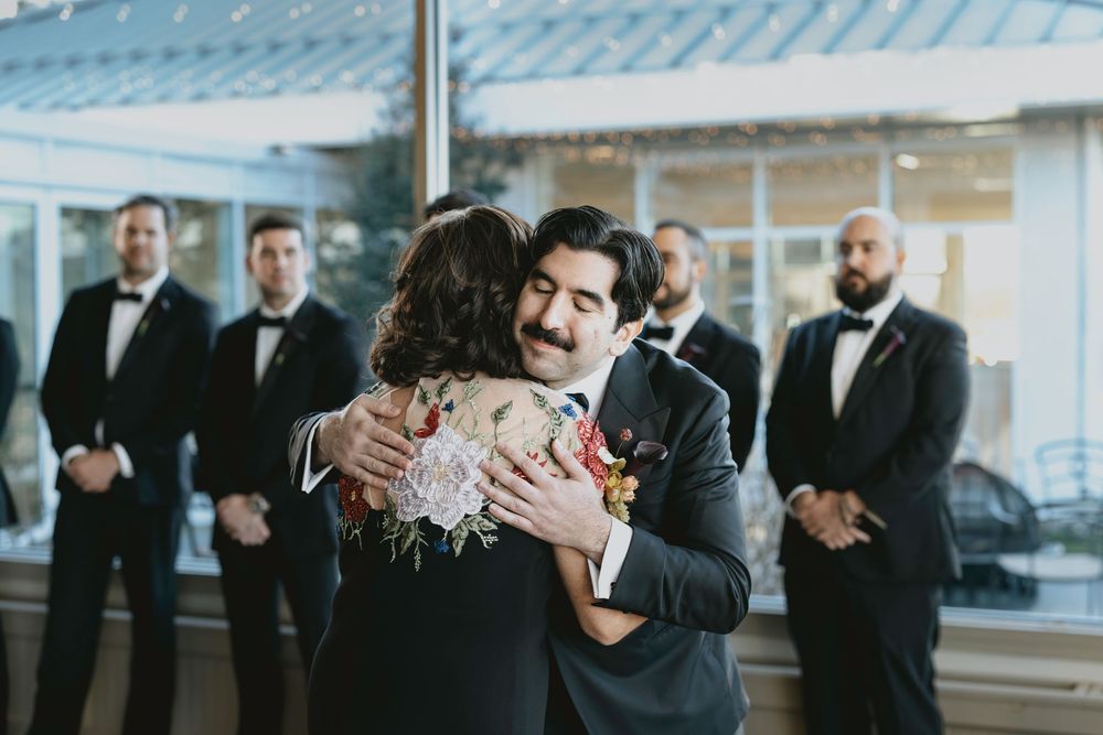Wedding guests and wedding party members gather for celebratory hugs and greetings at an indoor venue.