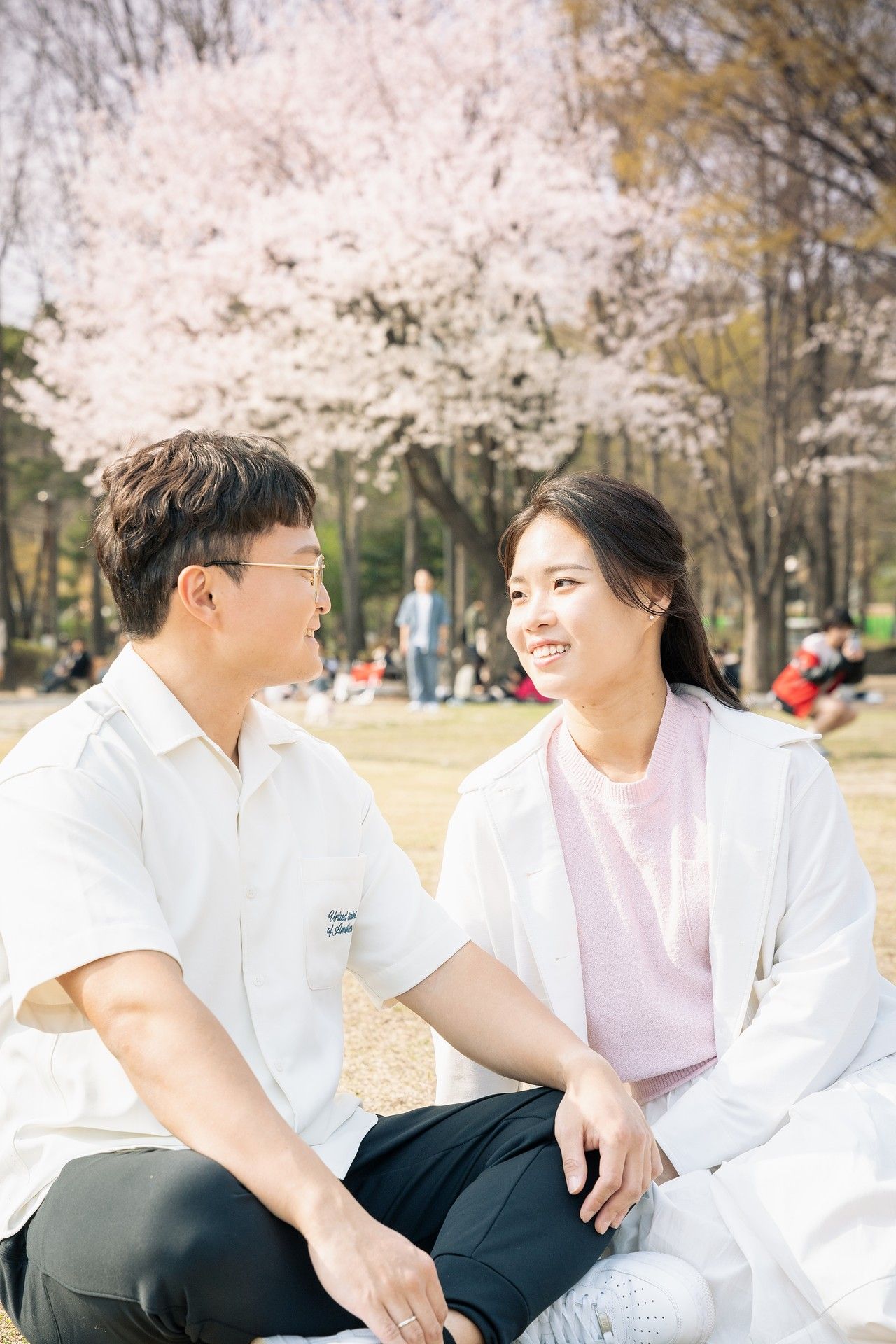 Two people sitting together outdoors under blooming cherry blossom trees in spring wearing casual white clothing.