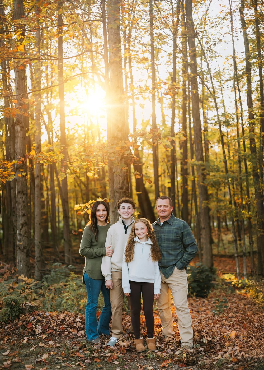 Autumn family photoshoot in a sunlit forest with golden leaves and tall trees.