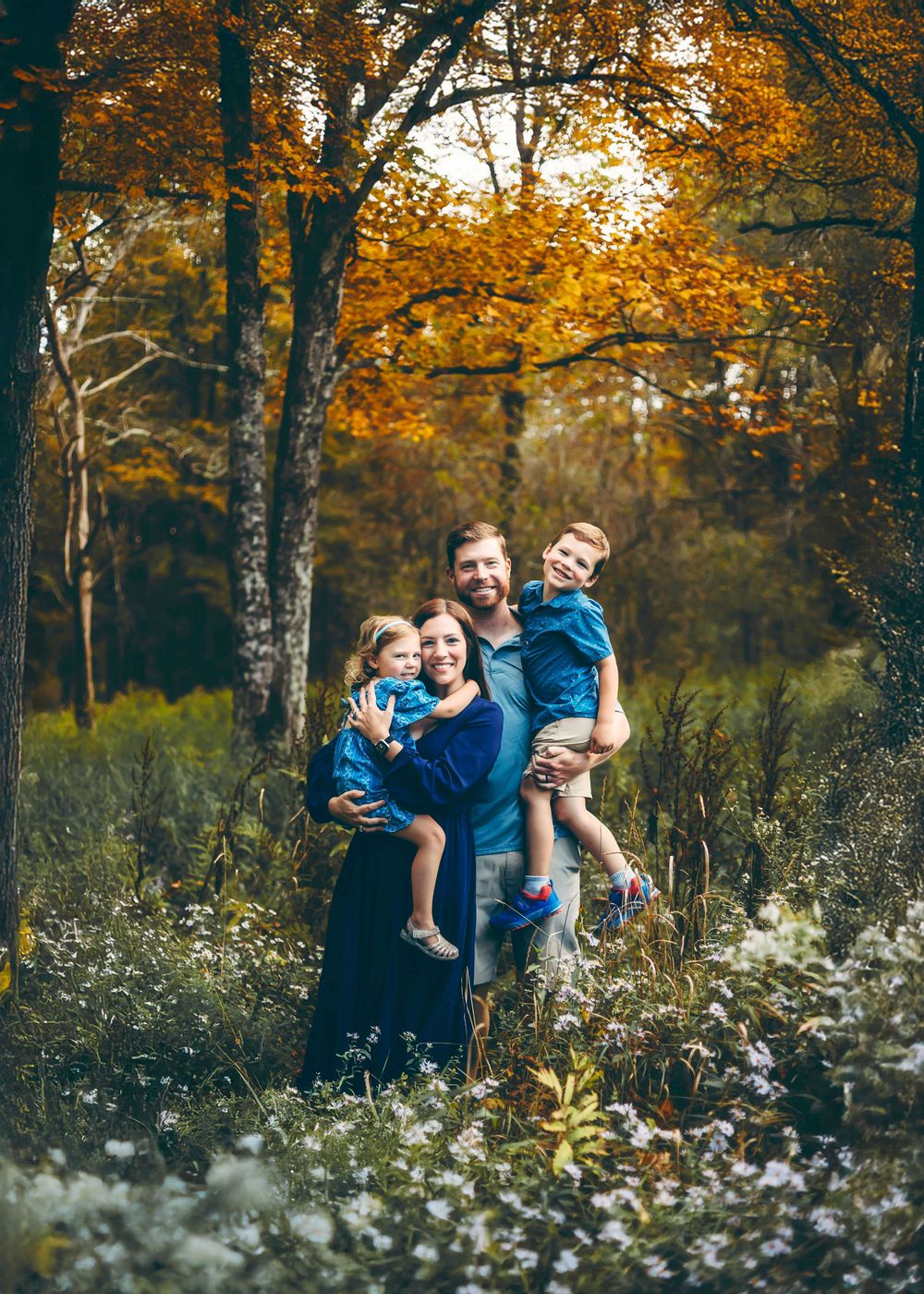 Family portrait in autumn forest with golden leaves and white wildflowers in foreground.