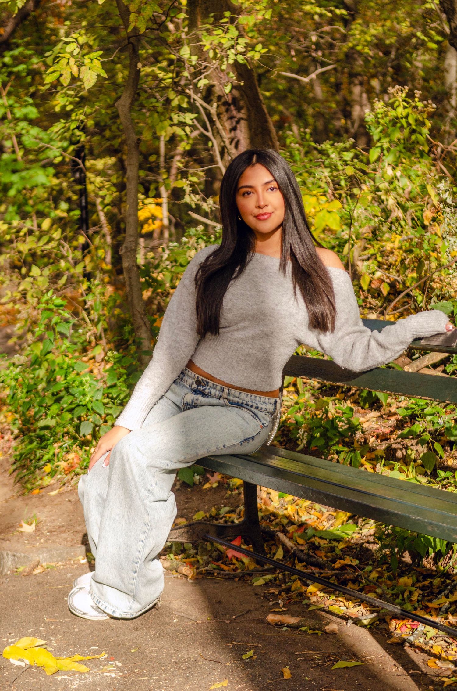 A woman in a gray sweater and white pants poses on a wooden bench in an autumn park setting with fallen leaves.