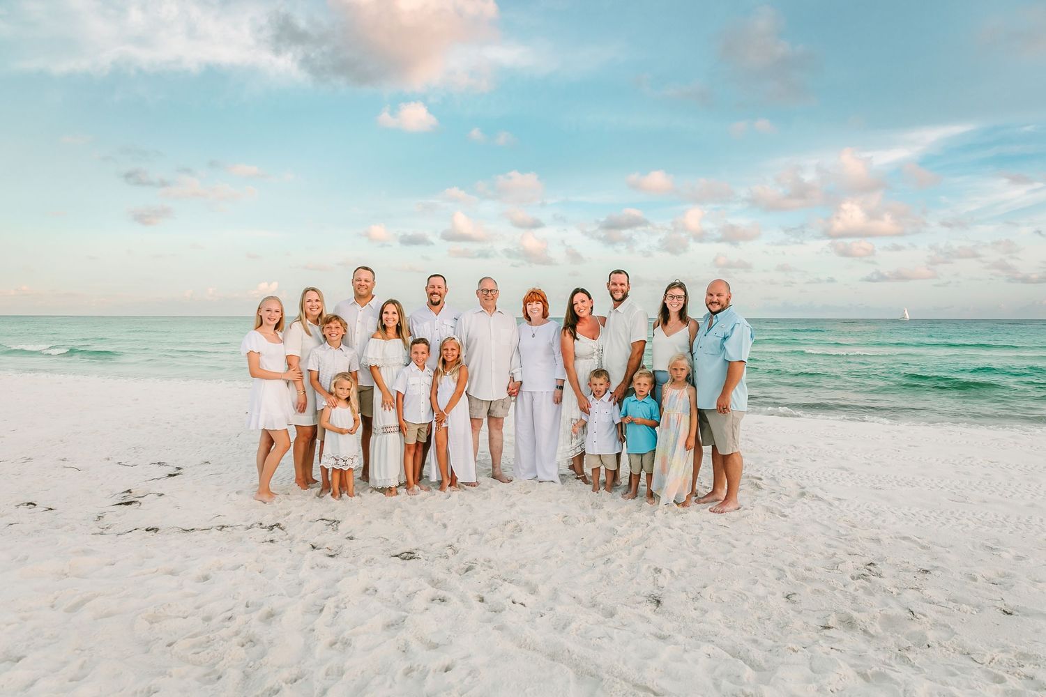 A group wearing white outfits poses together on a sunny beach with turquoise ocean waves in the background.