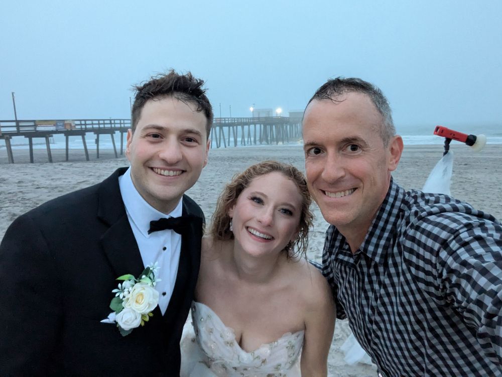 A group of three people pose for a selfie on the beach with a pier visible in the background at dusk.