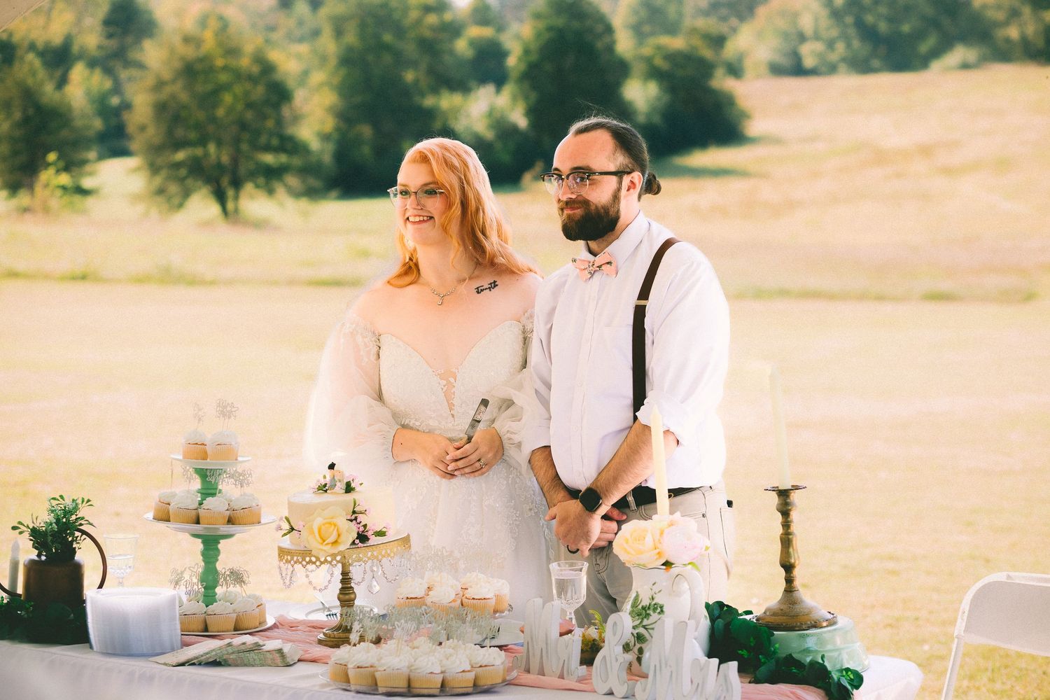 Elegant outdoor wedding reception table settings with white linens and floral centerpieces in a golden field at sunset.