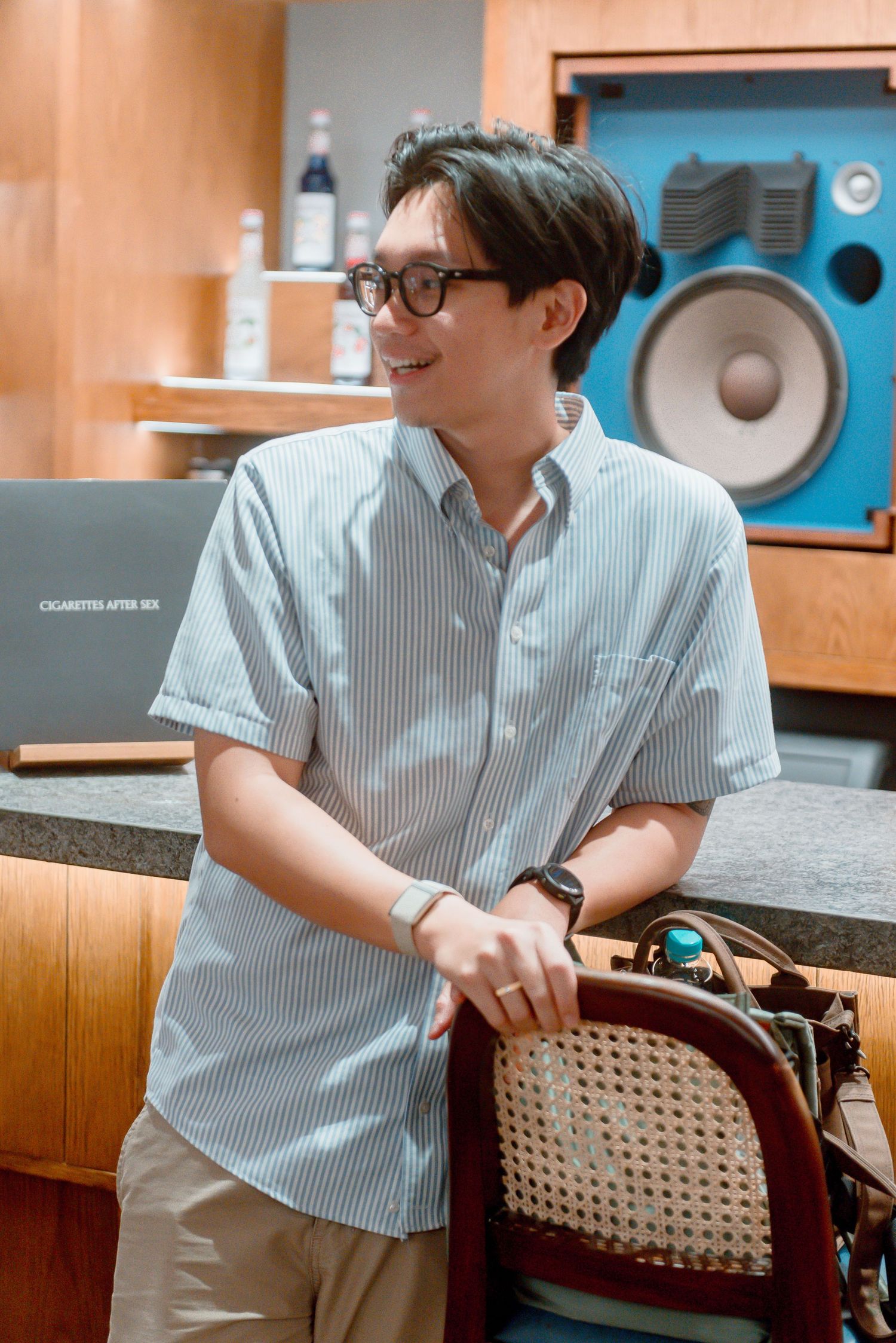 A person wearing glasses and a striped shirt sits in a woven chair at a recording studio.