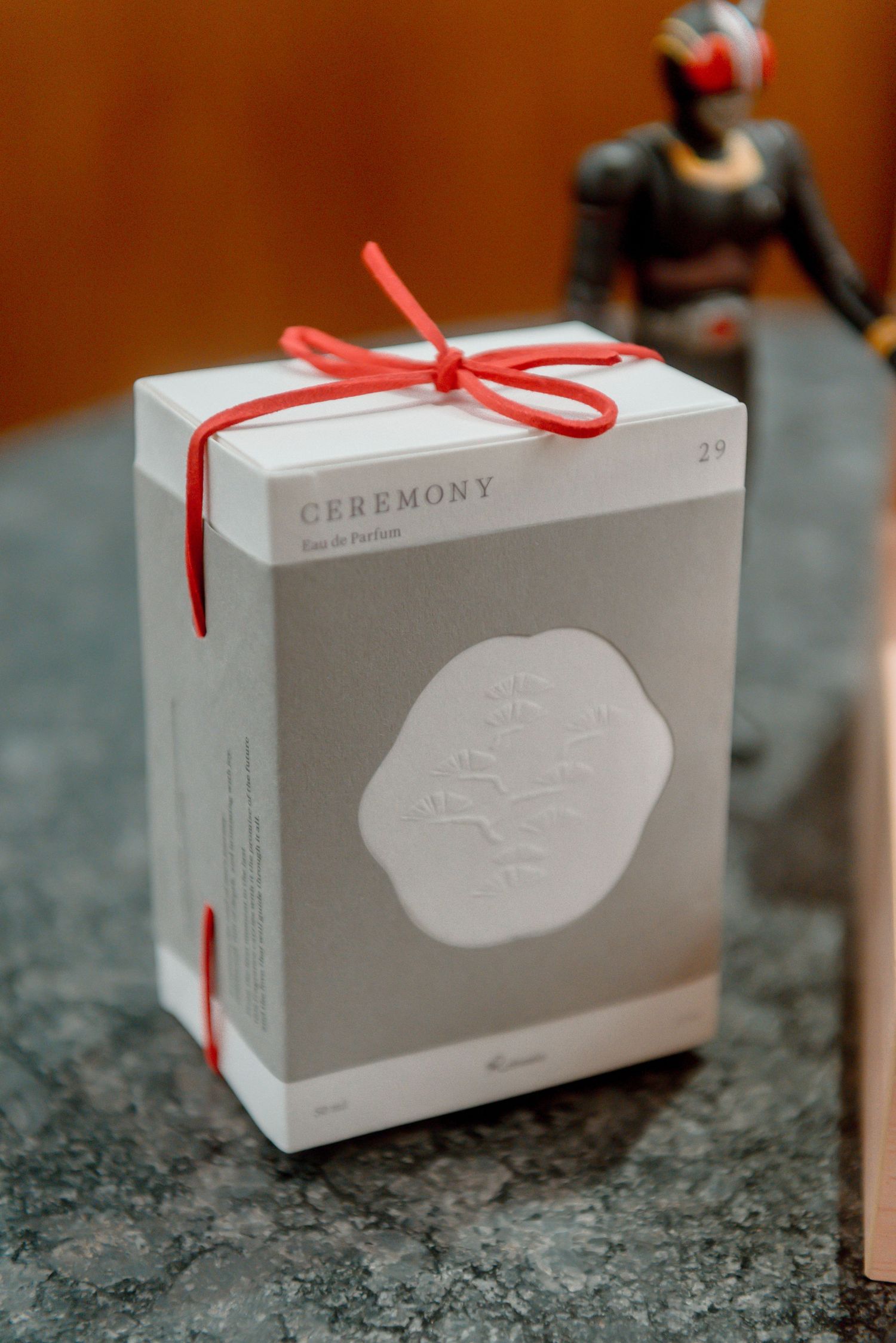 A white gift box with 'Ceremony' branding tied with a red ribbon bow sits on a gray marble surface.