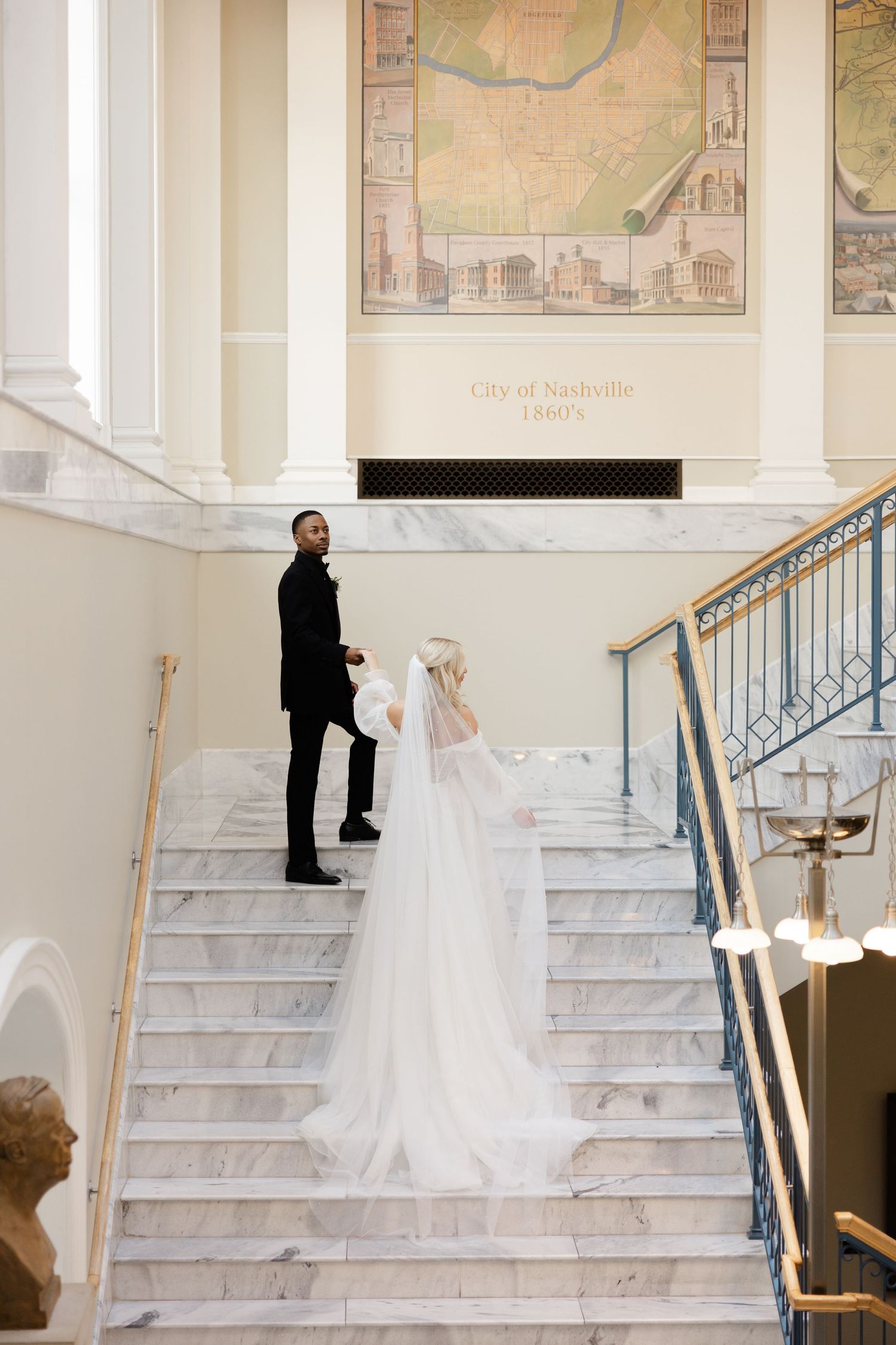 A couple in wedding attire stands on an elegant marble staircase with vintage maps decorating the wall behind them.