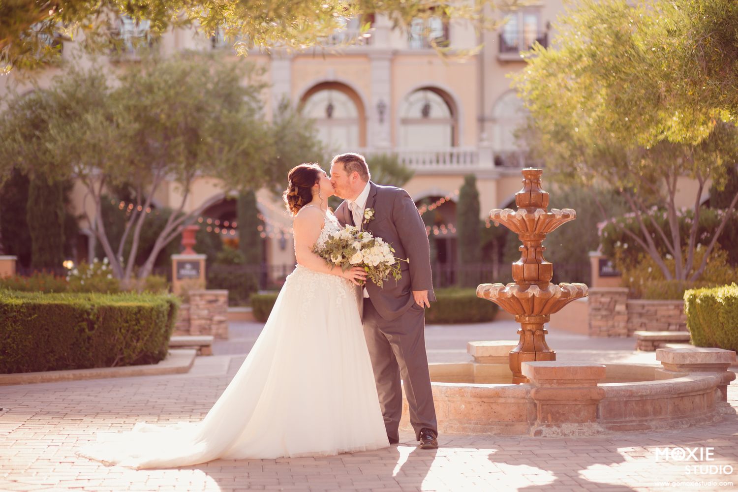 Bride and groom share a romantic kiss near a stone fountain in an elegant outdoor courtyard with lush greenery at Hilton Lake Las Vegas.