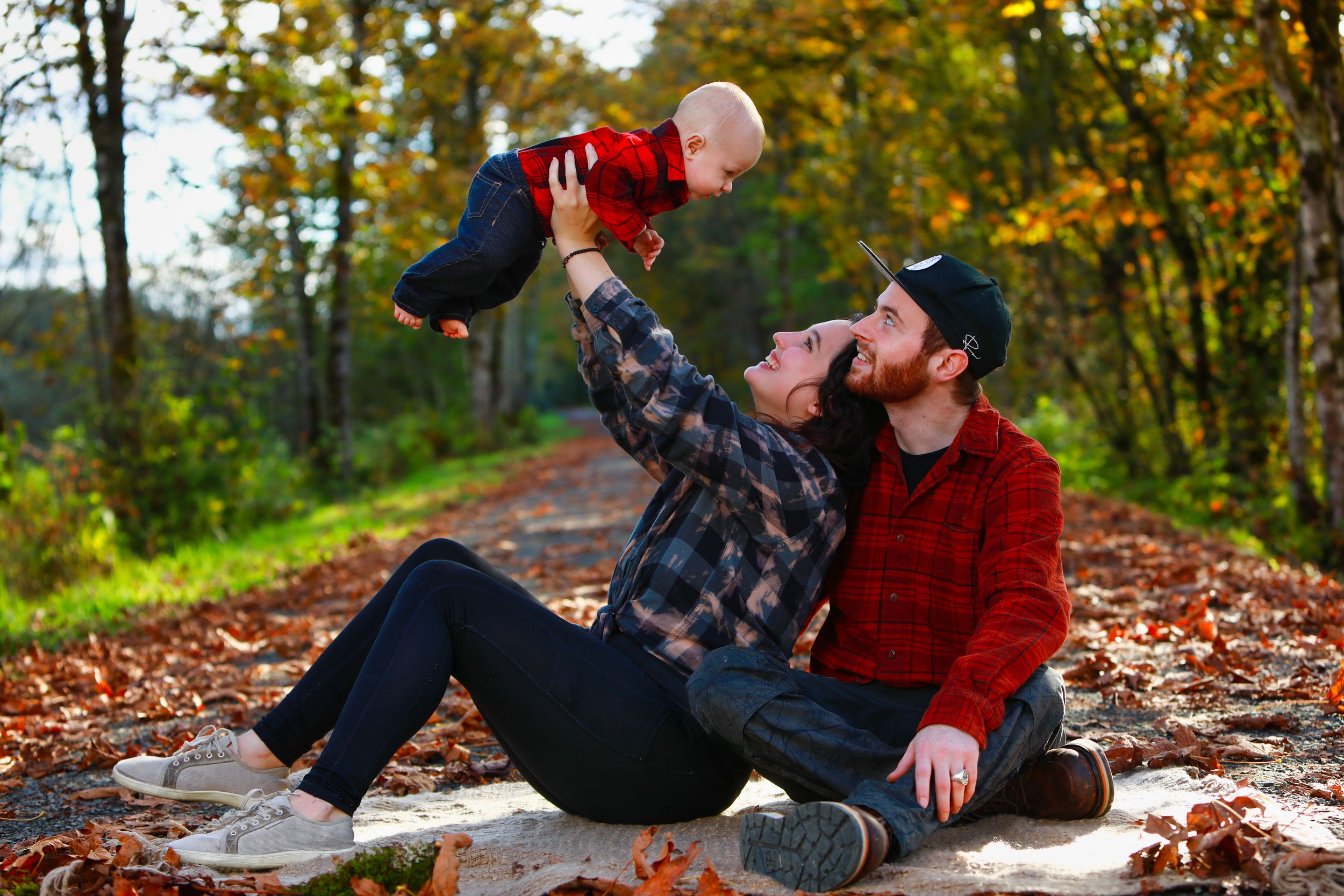 Two people in plaid shirts share a joyful moment while sitting on fallen autumn leaves in a forest setting.