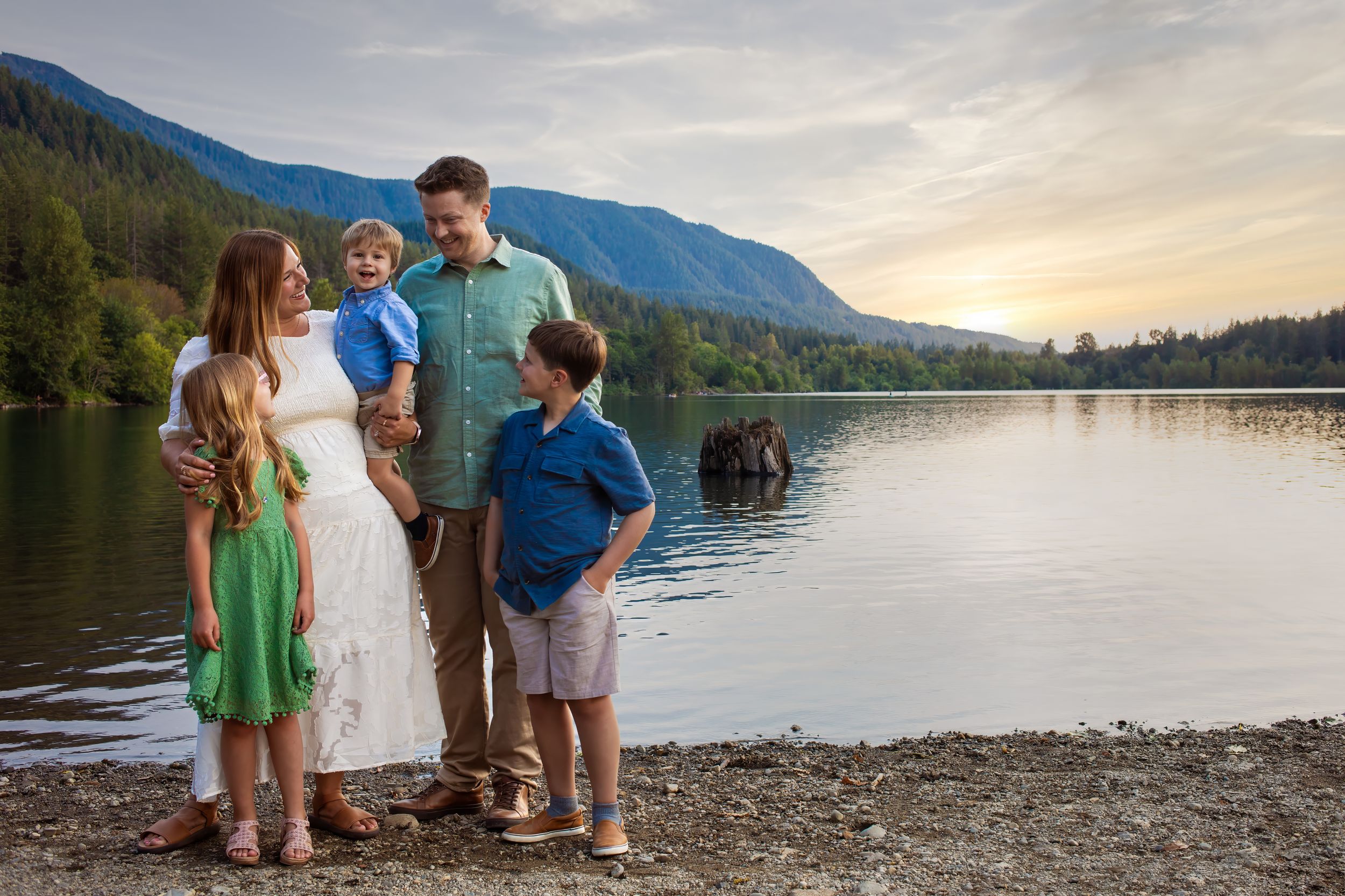 A family enjoys a serene sunset moment together on the rocky shores of a mountain lake with calm waters.
