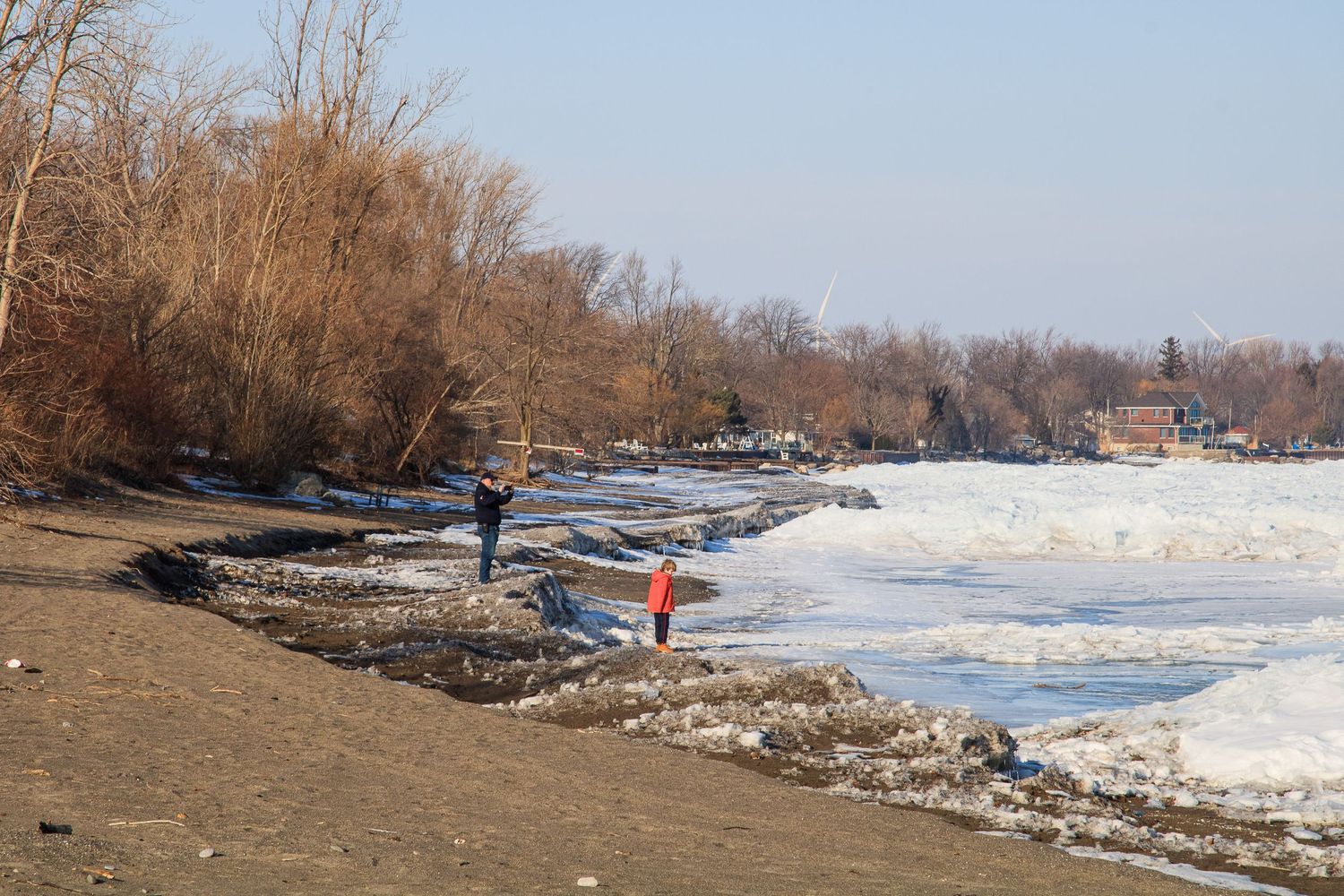 Winter landscape showing snow-covered riverbank with bare trees and debris along the shoreline in natural lighting.