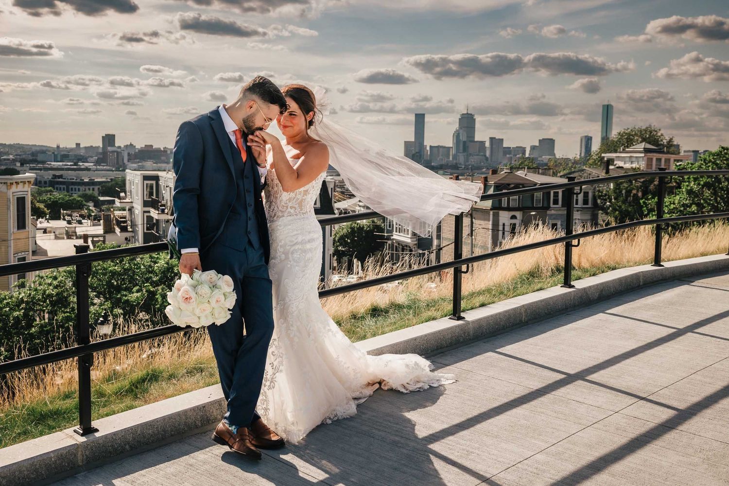 Bride lifting her veil as the couple shares a moment during wedding portraits at Dorchester Heights with the Boston skyline behind them