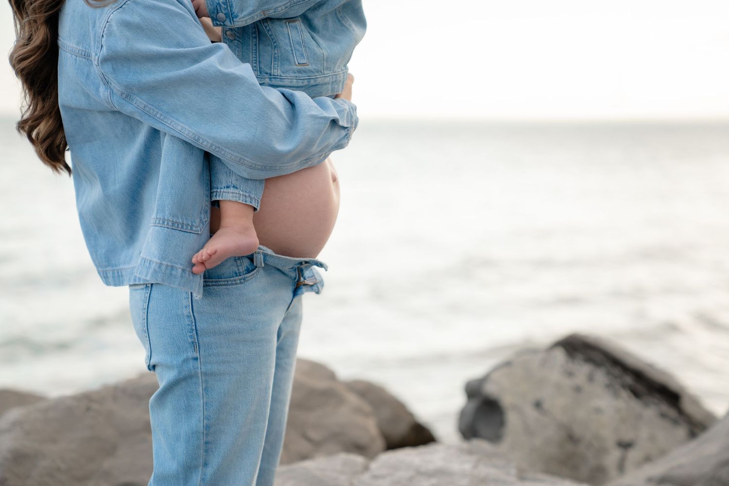Person wearing light blue denim outfit stands on rocky beach shoreline at sunset.