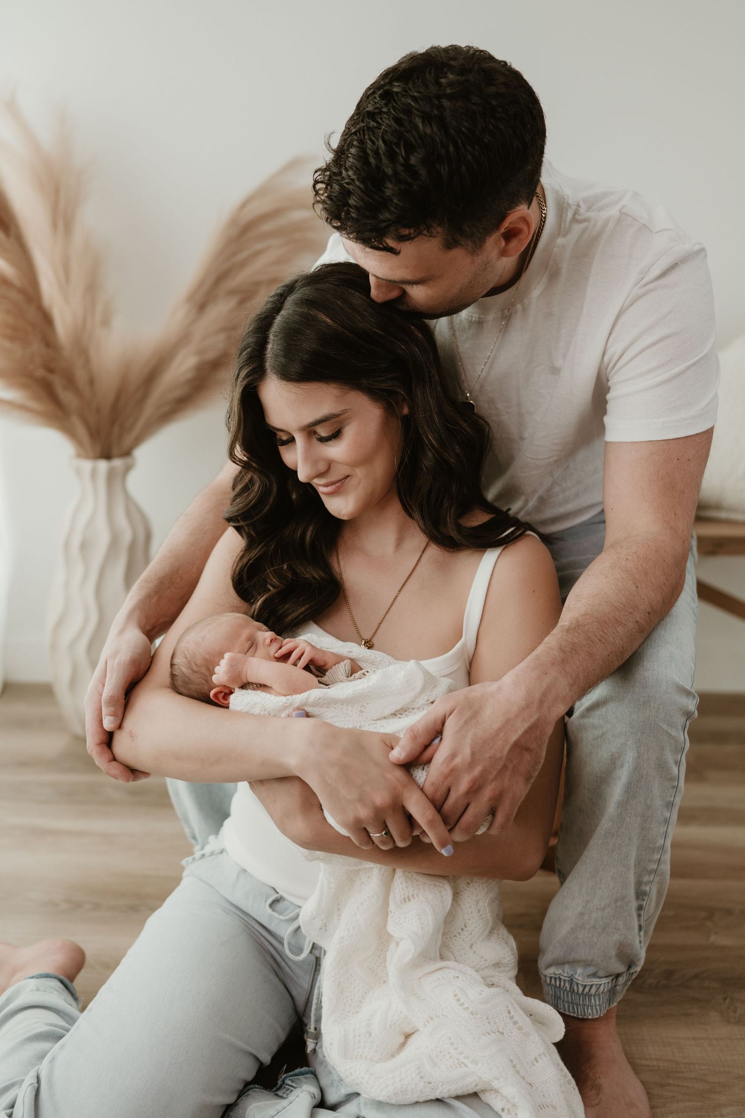 Pregnant woman in white top and jeans reclines on a chaise lounge, cradling her bare baby bump near pampas grass decor.