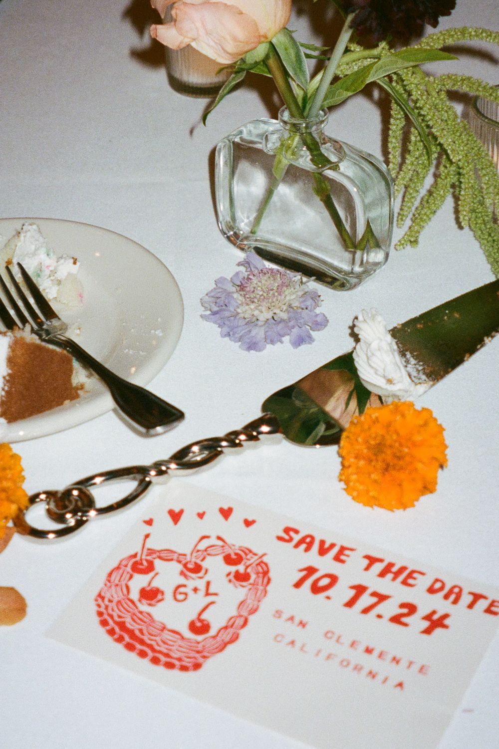 A save the date card with red hearts and flowers sits on a table with dessert plates and floral decorations.