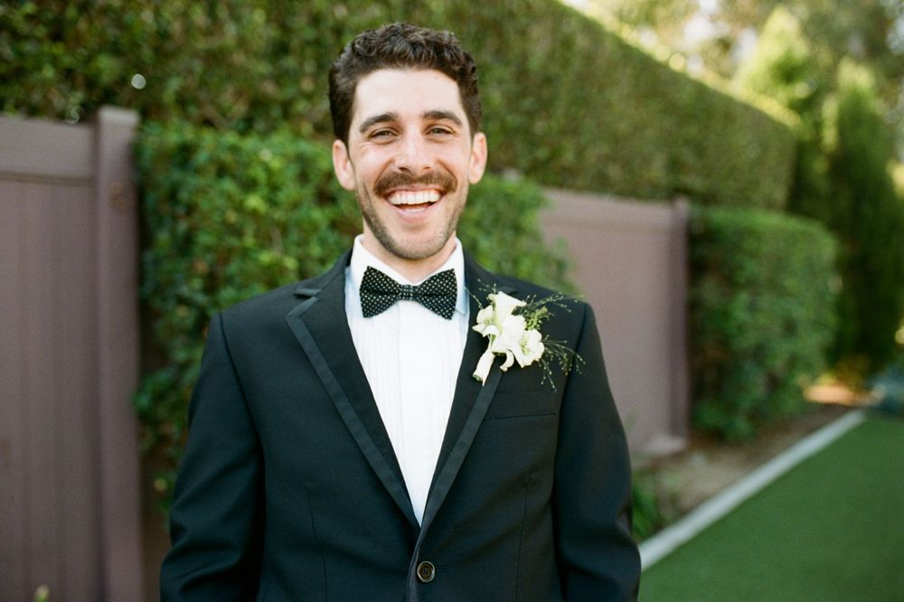 A groom in a black tuxedo and bowtie smiles warmly while standing outdoors during a wedding ceremony.