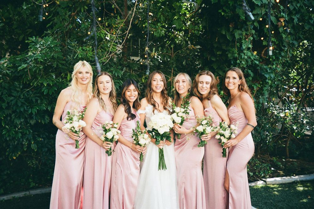 Bridesmaids in blush pink dresses pose together in an outdoor garden setting for wedding photos.