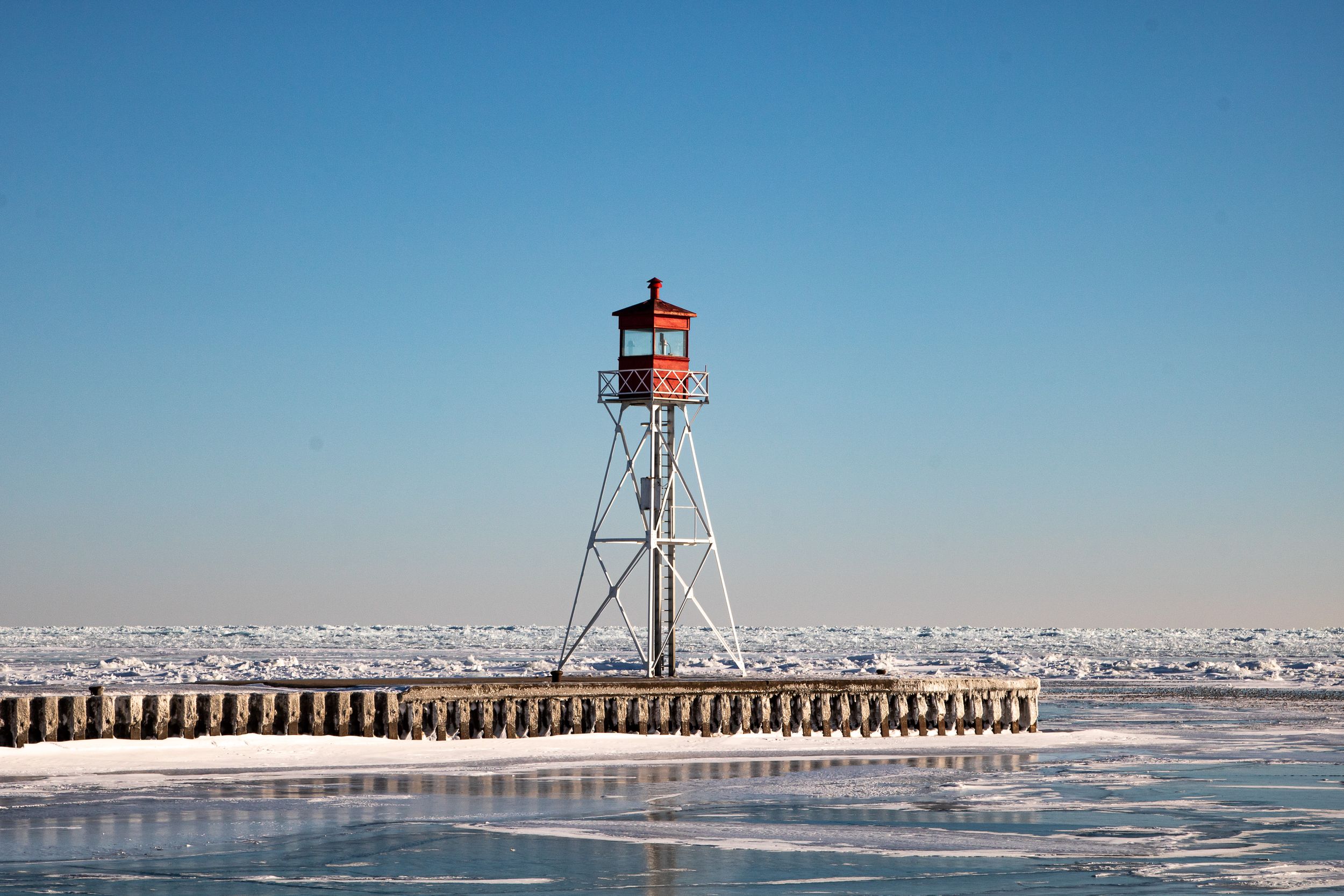 A small lighthouse tower stands on a snowy breakwater pier against a clear blue winter sky.