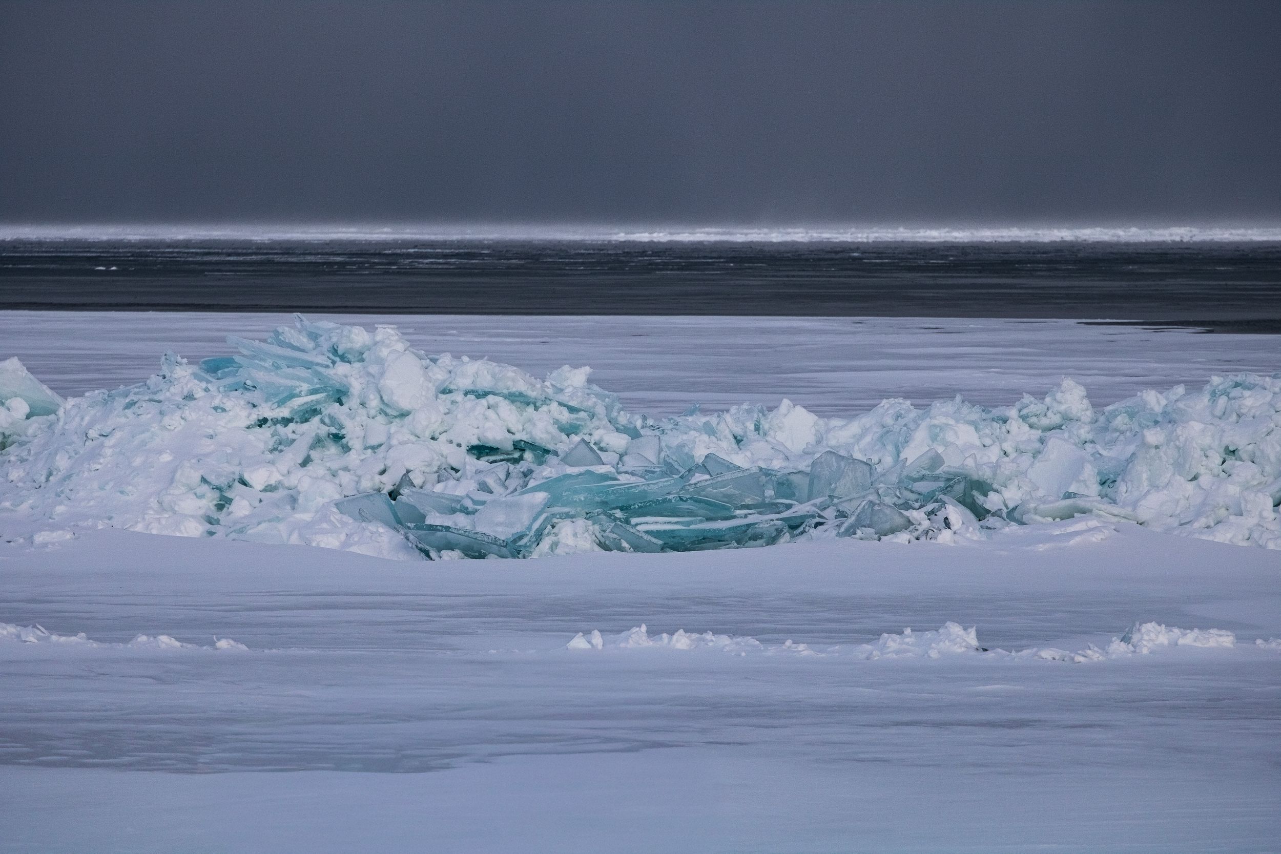 Jagged turquoise ice formations contrast against a snowy Arctic landscape under a moody gray sky.