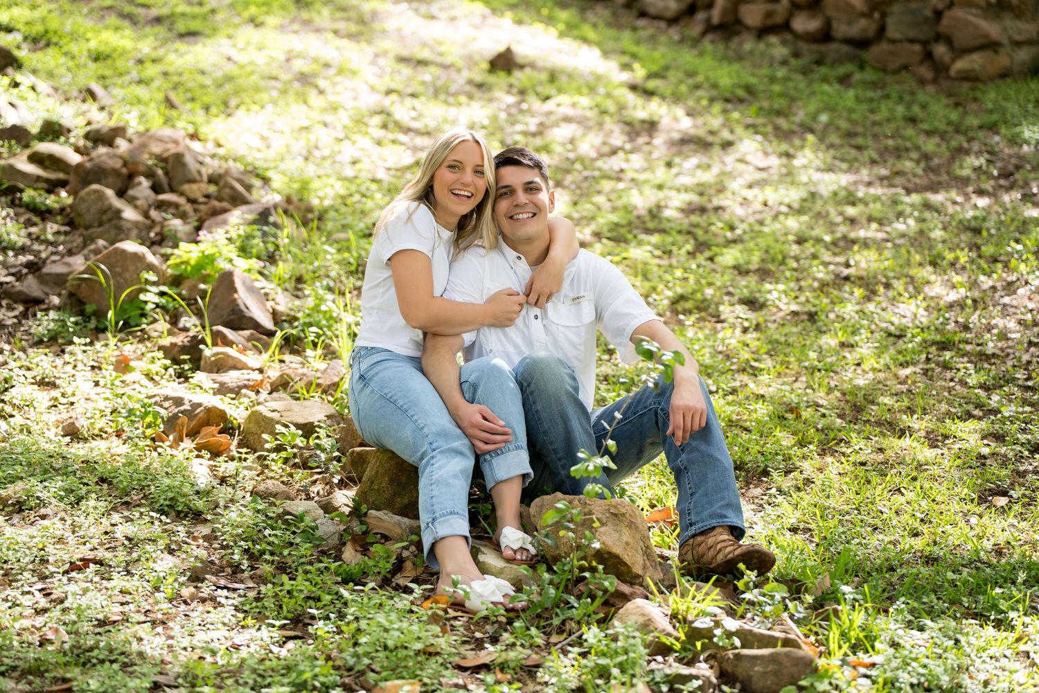A smiling couple sits together on a log in a lush green park, wearing casual white tops and jeans.