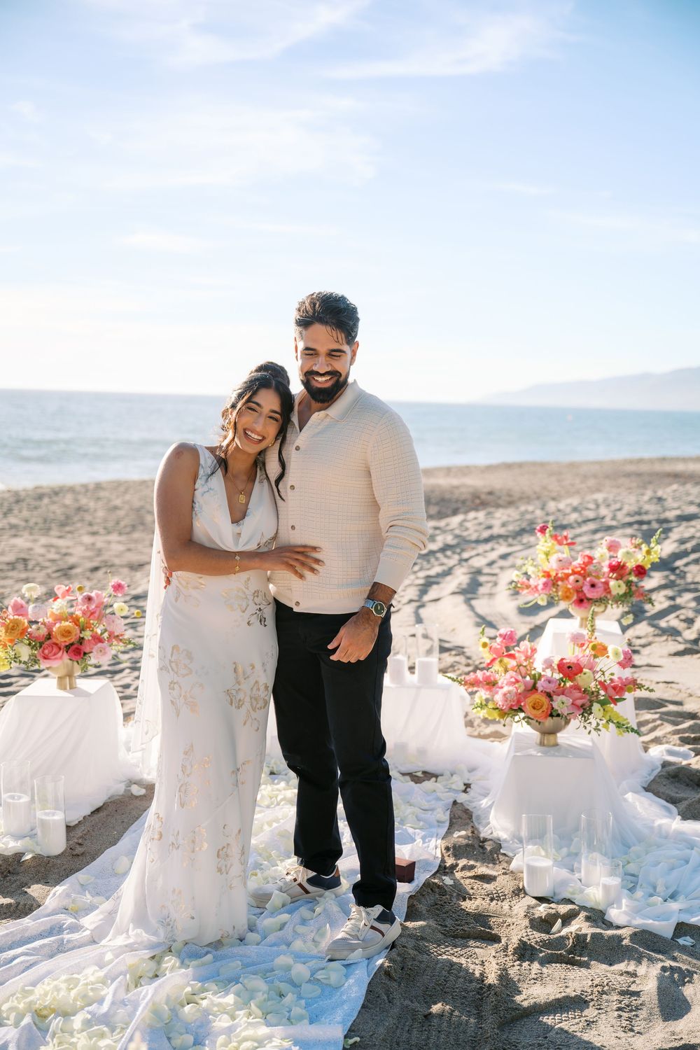 A romantic beach wedding setup with a couple embracing by colorful floral arrangements on the sand at sunset.