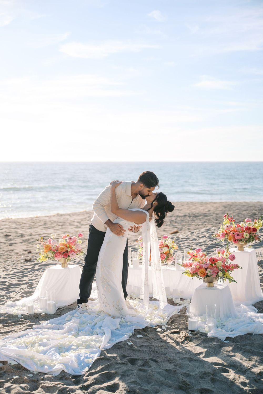 A romantic beach wedding scene with a couple sharing an intimate moment beside elegant table settings and coral flowers.