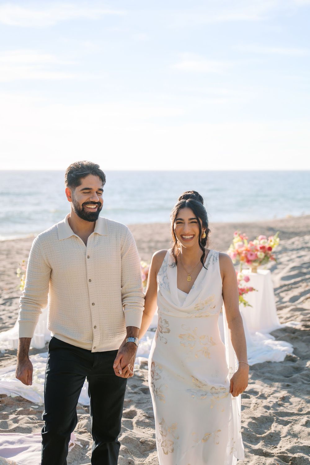 A joyful couple in white attire laughing together on a sandy beach during their wedding celebration.