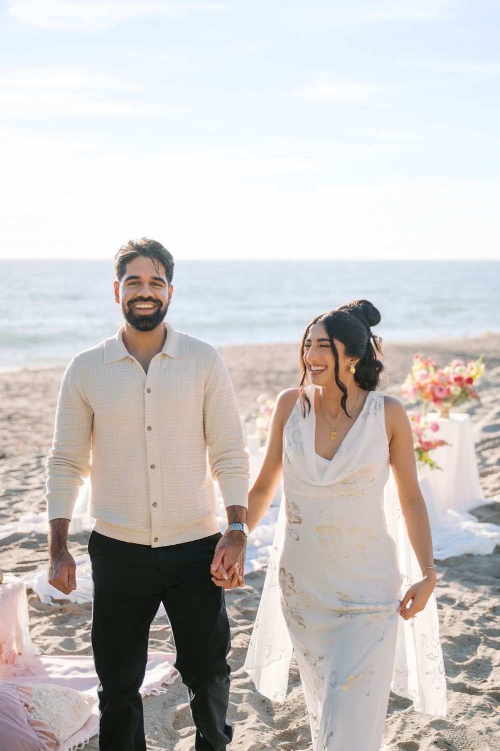 A happy couple laughs and walks together on a sunny beach during a romantic photoshoot.