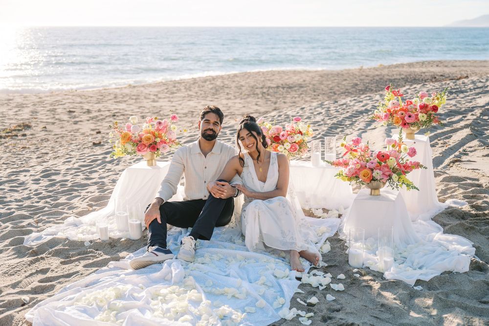 A romantic beach wedding celebration with white linens and pink floral arrangements along the sandy shore at sunset.