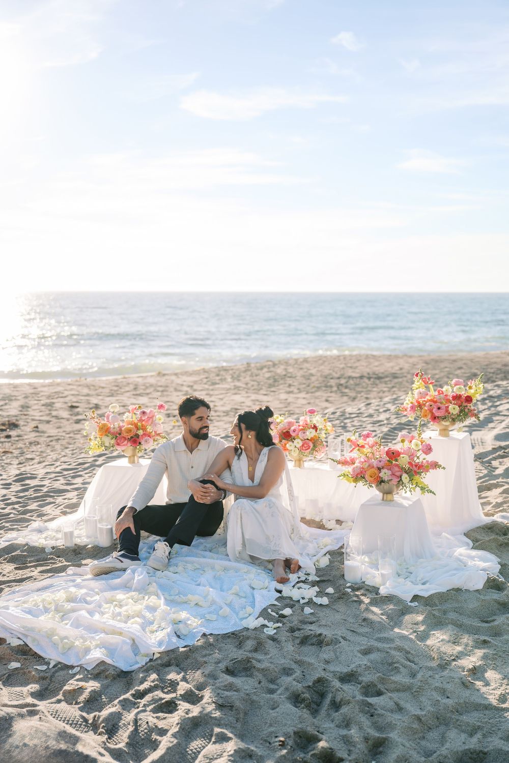 Romantic beach picnic setup with white linens and colorful floral arrangements on sandy shore at sunset.