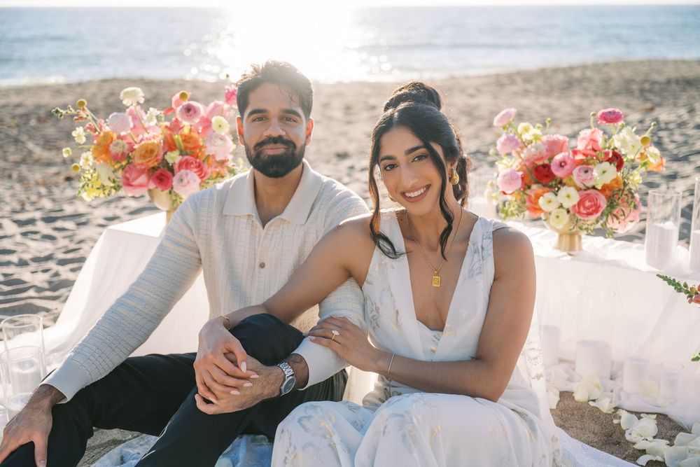 A couple sits on a sandy beach with pink and coral floral arrangements during a romantic seaside wedding celebration.