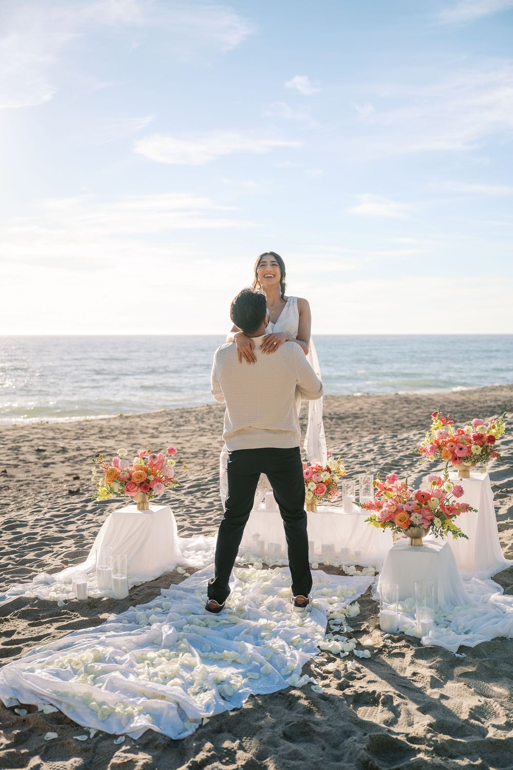 A romantic beach wedding setup with white linens and coral floral arrangements on tables against a coastal backdrop.