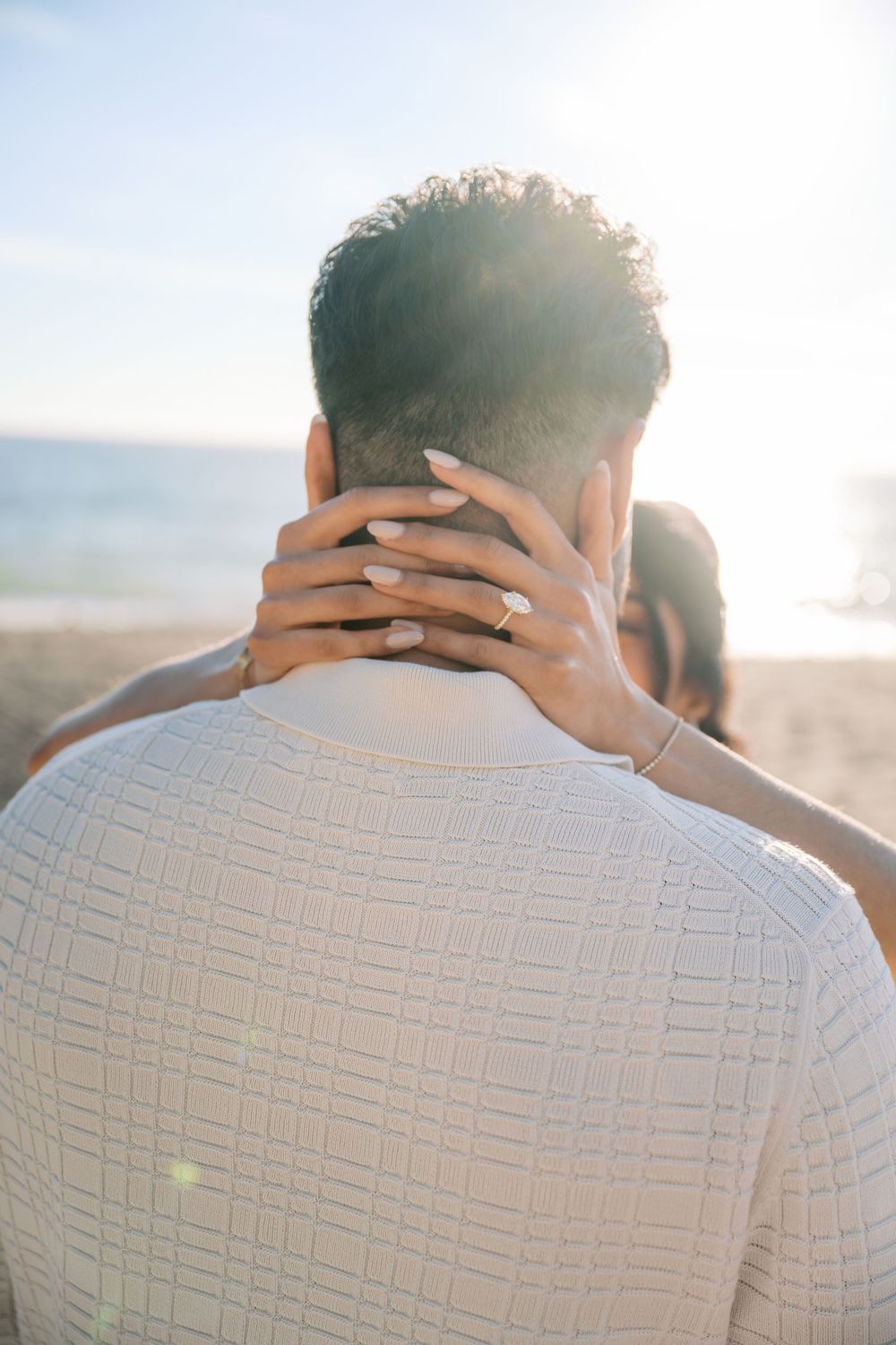 A romantic couple embraces on a sunlit beach while wearing a white textured shirt.