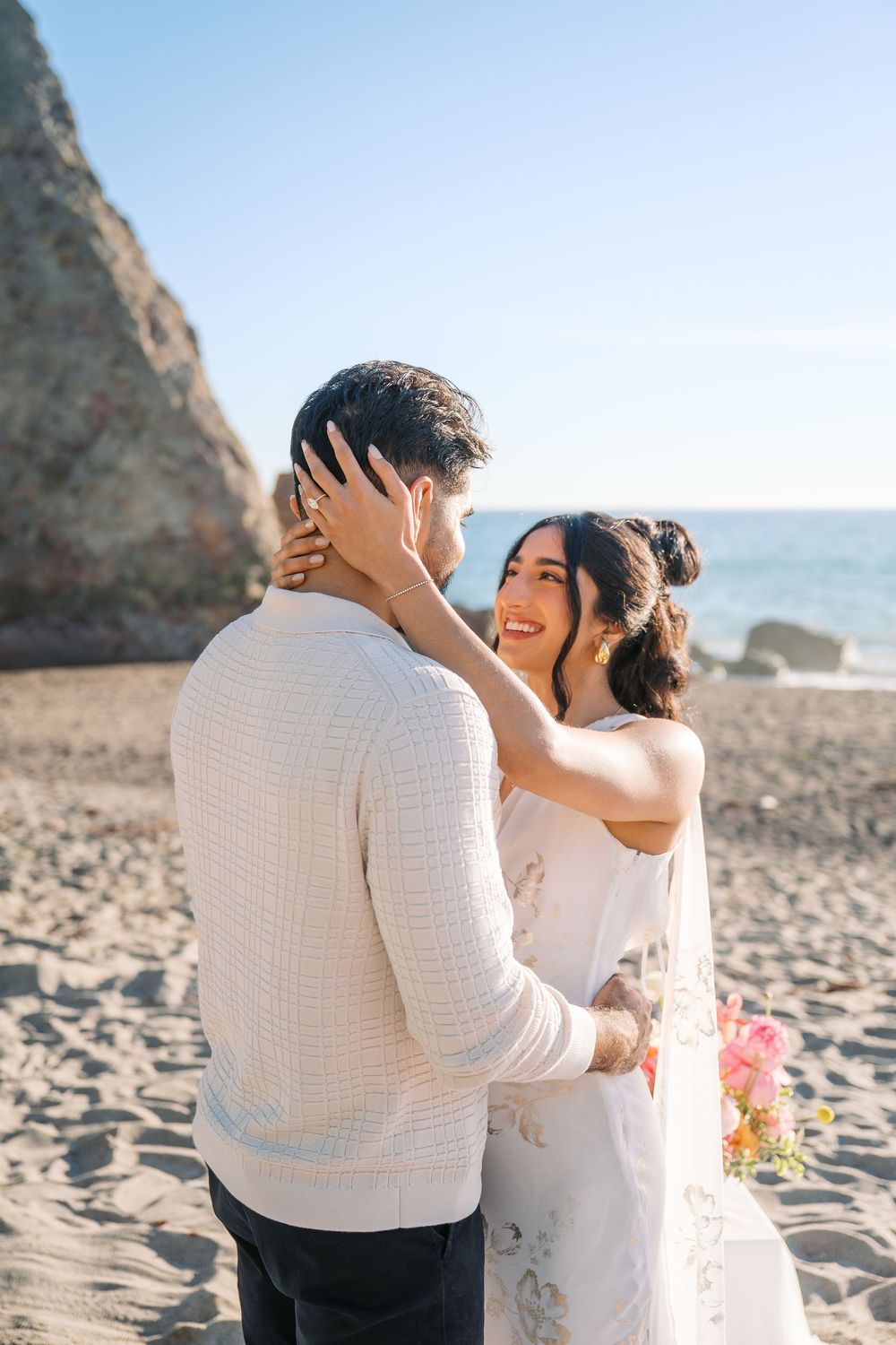 A couple shares a romantic moment on a sunny beach with rocky cliffs in the background.