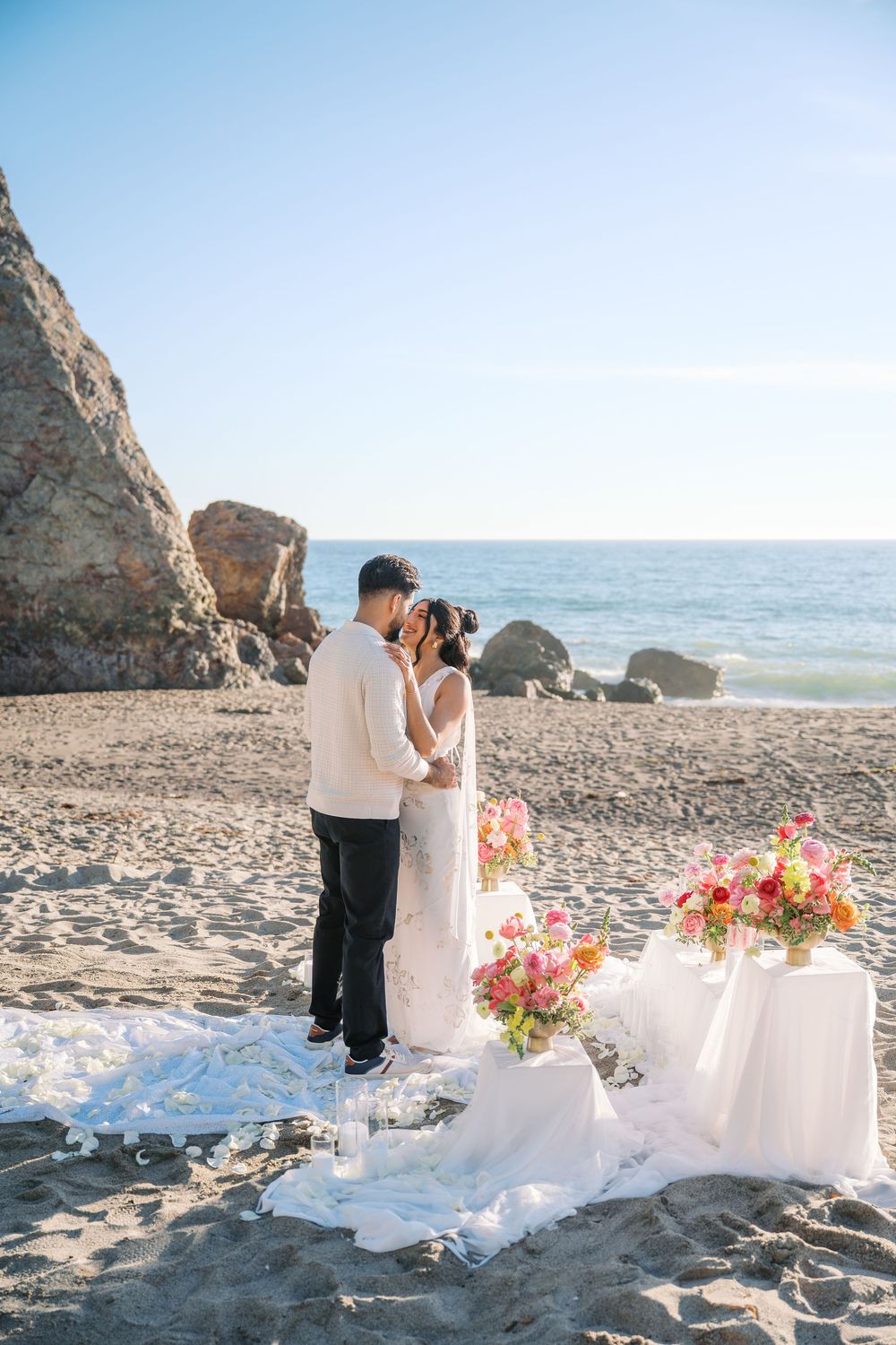 Romantic beach wedding setup with white linens and colorful floral arrangements along a rocky coastline at sunset.