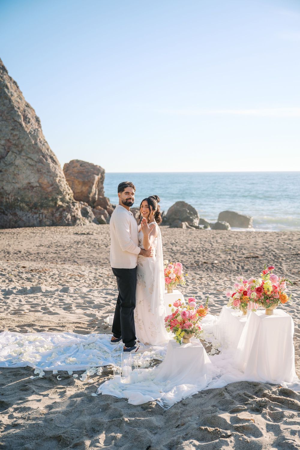 A romantic beach wedding setup with white linens and coral floral arrangements against rocky coastal cliffs at sunset.