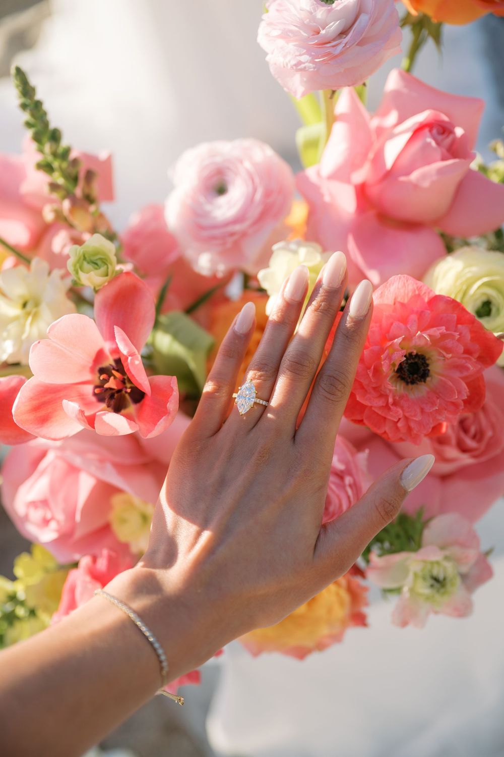 Close-up of hands displaying wedding rings among pink and coral-colored floral arrangements.