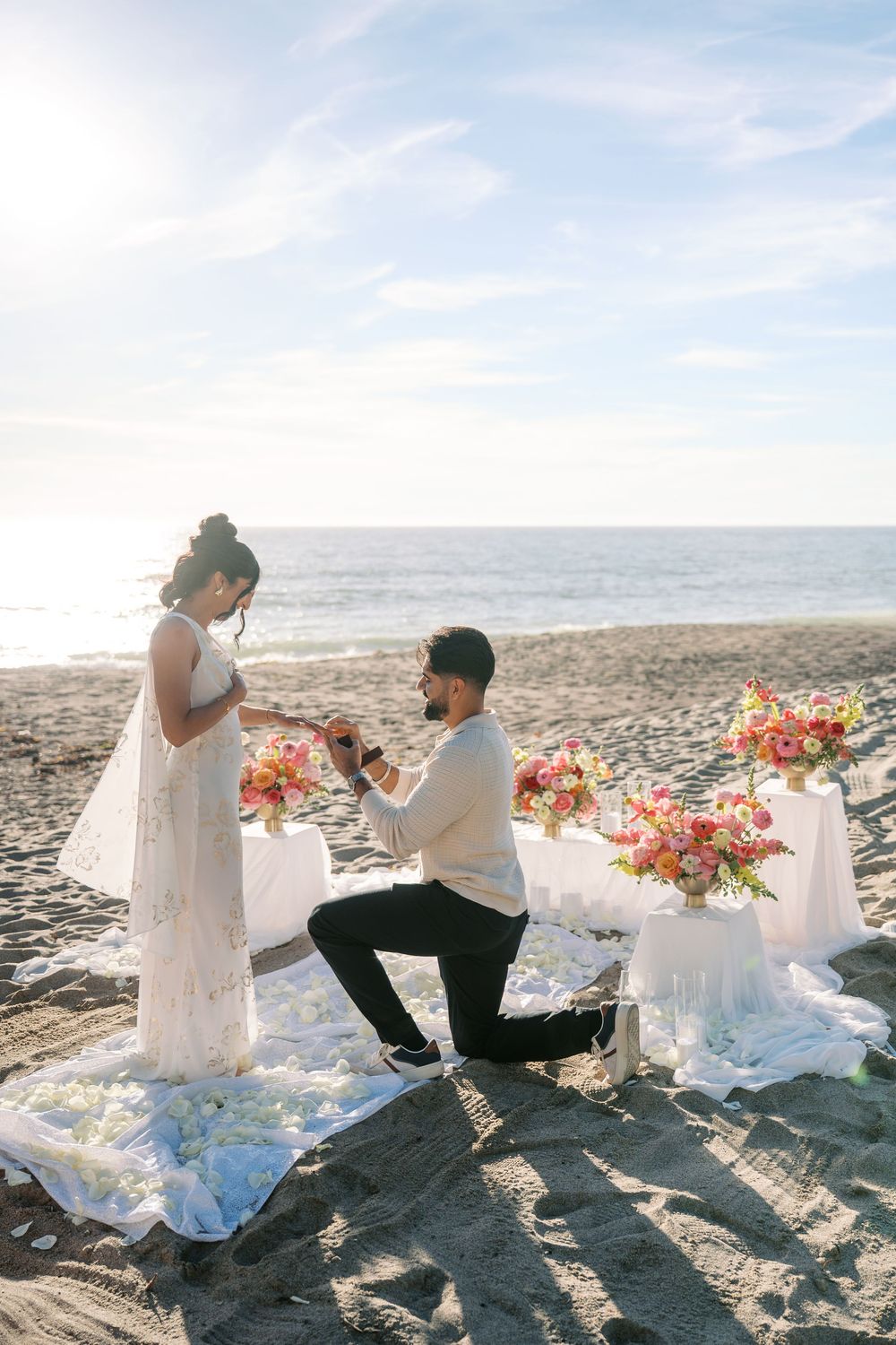 A romantic beach proposal scene with flowers and decorations at sunset along the shoreline.
